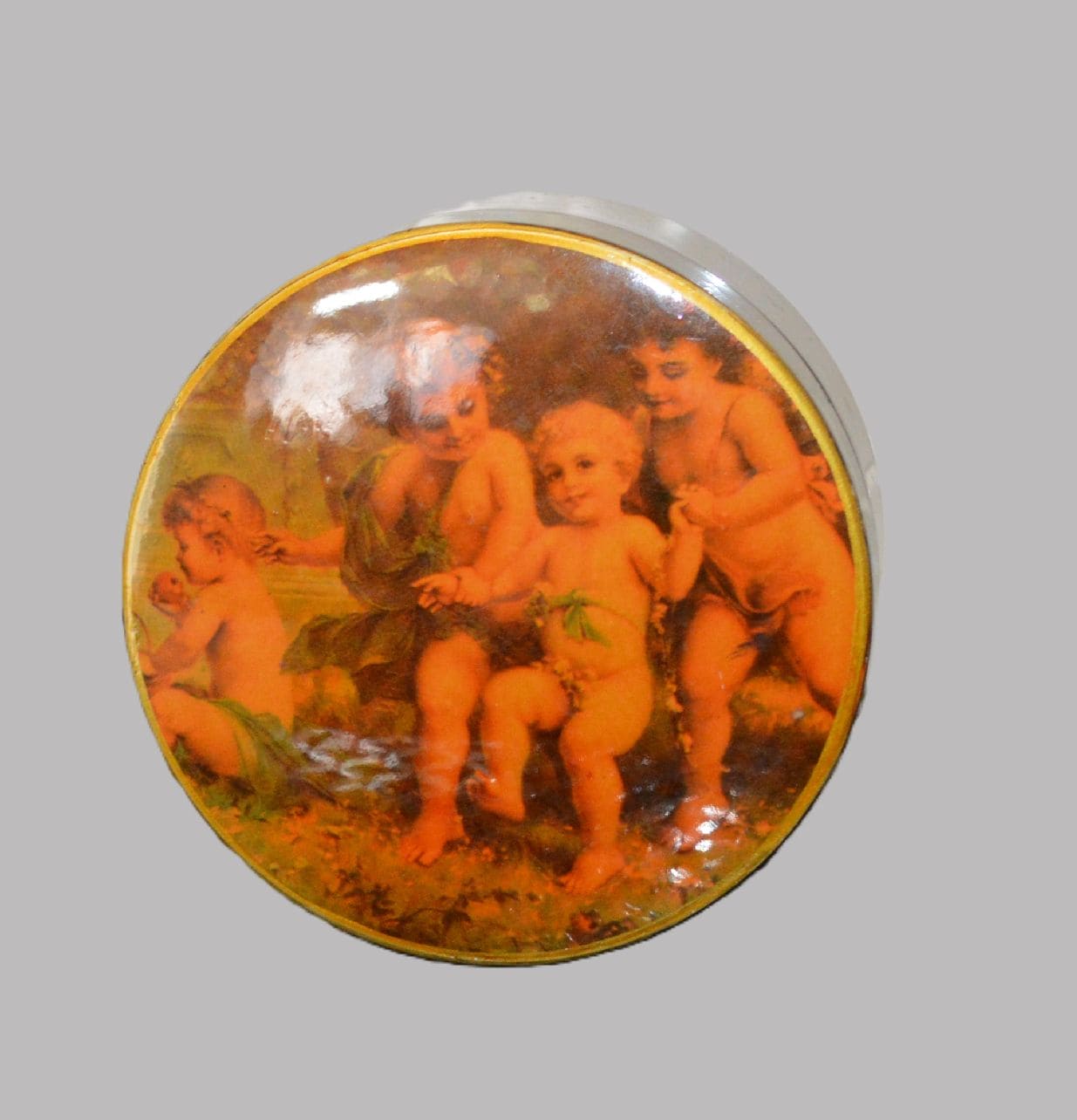 A round decorative box with a lid featuring a classical painting of four cherubic children. The children are depicted in an outdoor setting, with foliage in the background. They are unclothed with some greenery draped around them. The box itself appears to be dark-colored, possibly black, with the artwork in warm tones on the top of the lid. The box is placed on a green and red surface.