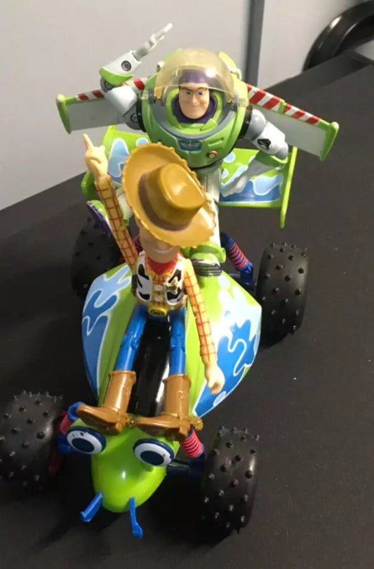 Toy Story action figures of Woody and Buzz Lightyear riding on a free-wheeling RC car. Buzz is positioned at the back with his wings extended, while Woody sits at the front pointing forward. The RC car features a green and blue design with large black wheels