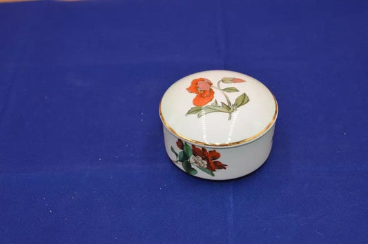 A small, round ceramic box with a white surface, decorated with delicate floral illustrations. The lid features a red flower with green leaves, and a gold trim lines the edge of the box. The box is set on a green background.